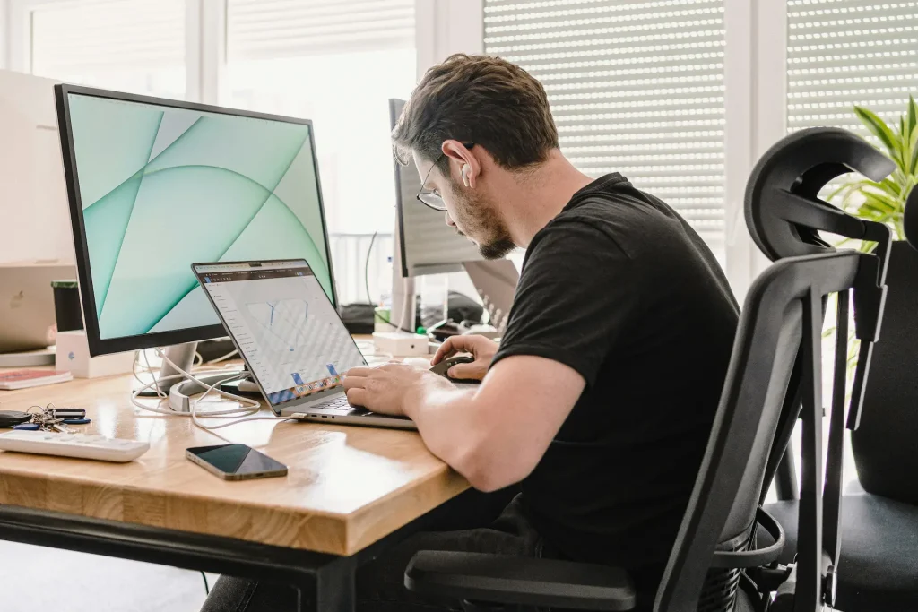 Développeur tapant sur un laptop, second écran branché, assis sur une chaise ergonomique près d’une fenêtre