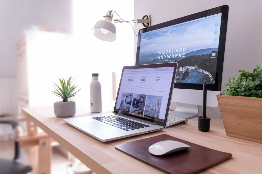 Bureau avec 2 ordinateurs, smartphone, souris sans fil, carnets et plante verte sur fond blanc.