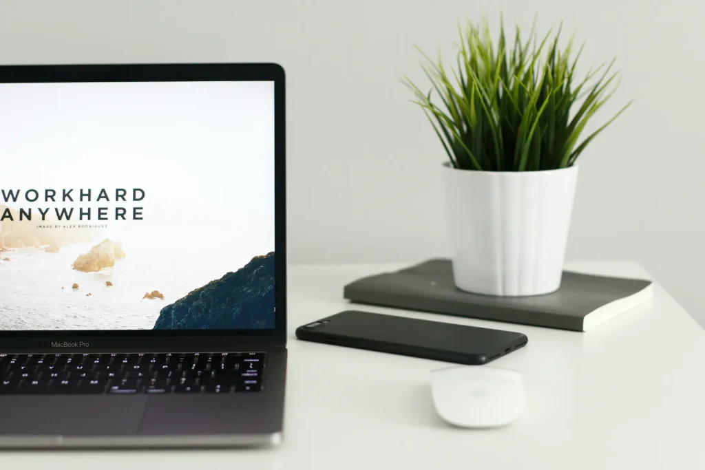 Bureau minimaliste avec ordinateur portable affichant “Work Hard Anywhere”, smartphone, souris sans fil, carnets et plante verte sur fond blanc.