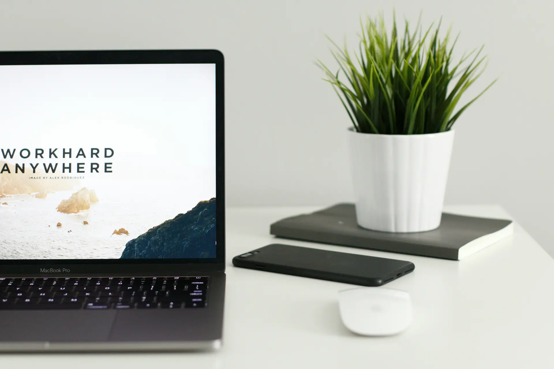 Bureau minimaliste avec ordinateur portable affichant “Work Hard Anywhere”, smartphone, souris sans fil, carnets et plante verte sur fond blanc.