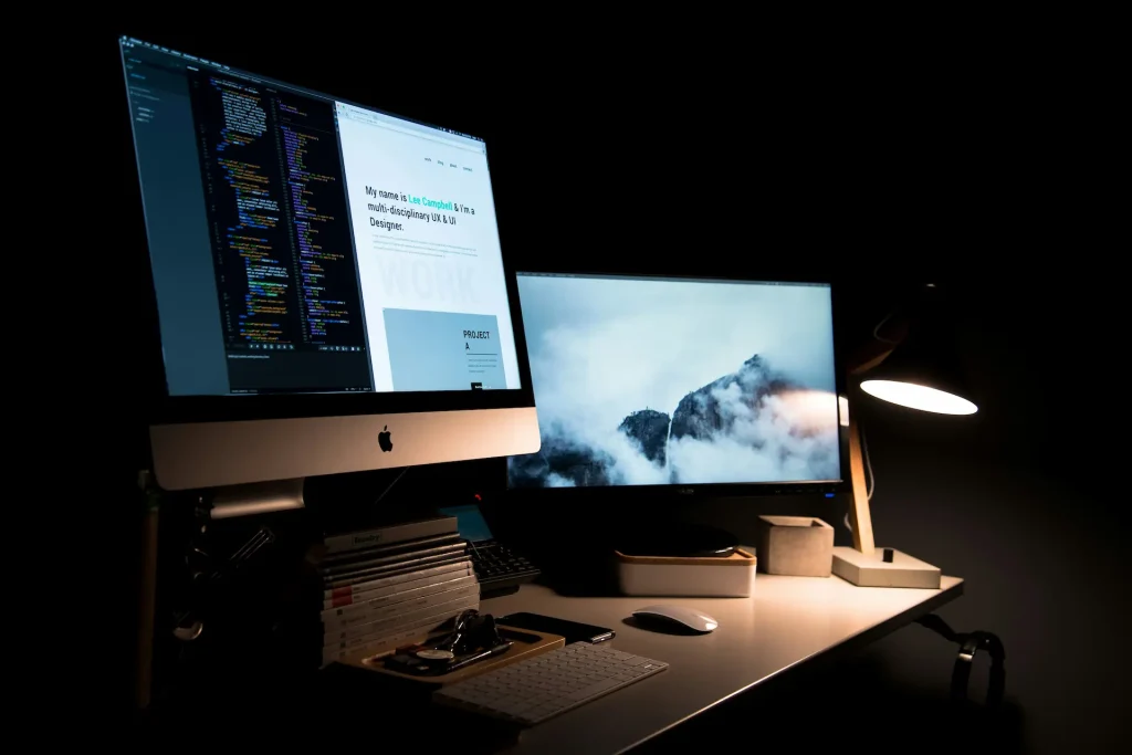 Espace de travail sombre avec deux écrans — code sur l’un, paysage montagneux sur l’autre, lampe de bureau