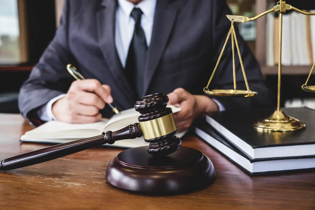 Maillet de juge et balance dorée sur un bureau, avec une personne en costume écrivant dans un carnet, symbolisant la justice et le droit.
