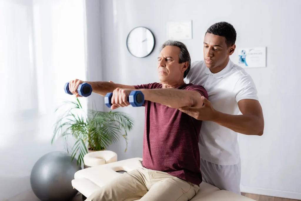 Un kinésithérapeute assiste un patient âgé qui soulève de petites haltères, bras tendus, lors d’un exercice de rééducation dans un cabinet lumineux.