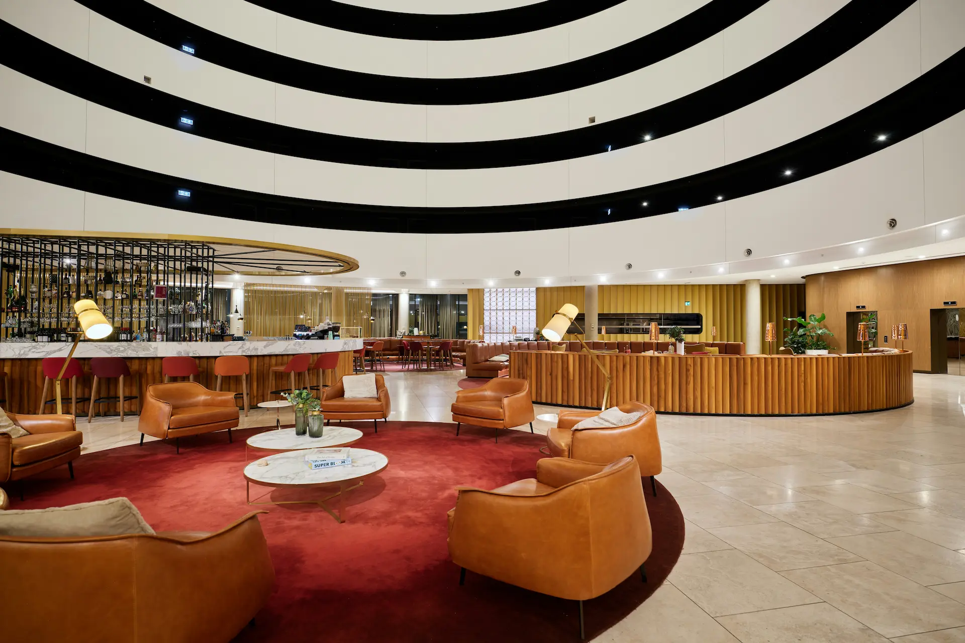Lobby d’hôtel design avec bar circulaire, fauteuils en cuir et grand tapis rouge, espace lounge lumineux et accueillant.